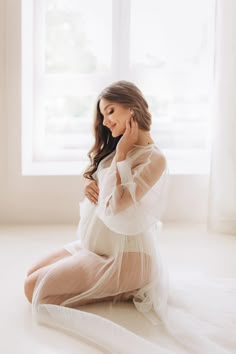 a pregnant woman sitting in front of a window wearing a white dress and holding her hands to her chest