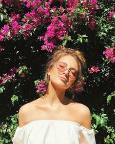 a woman wearing sunglasses standing in front of purple flowers with her head tilted to the side