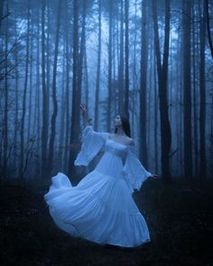 a woman in a white dress is standing in the woods with her arms spread out