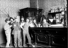 an old black and white photo of men standing in front of a bar with bottles on it