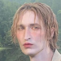 a man with blonde hair and blue eyes looks at the camera while standing in front of trees