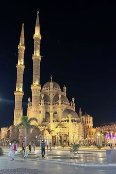 people are walking around in front of a large building at night with lights on it