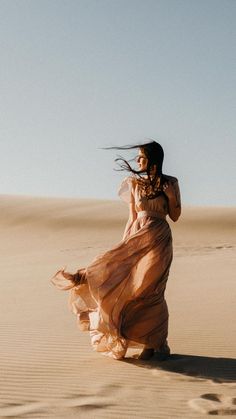 a woman in a long dress is walking through the desert with her hair blowing in the wind