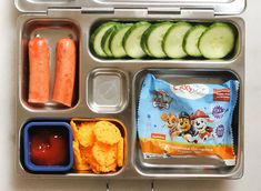 an open lunch box with vegetables, crackers, and snacks in it on a table