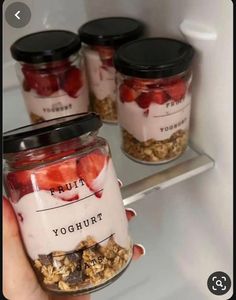 a person holding a jar filled with yogurt and strawberries in front of an open refrigerator door