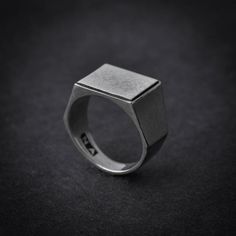 a silver ring with a square shaped design on the front and center, sitting on a black surface