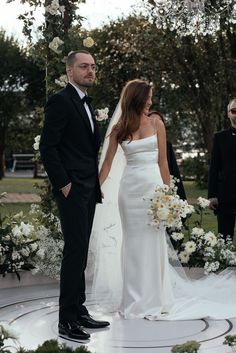 the bride and groom are standing in front of an arch with white flowers on it