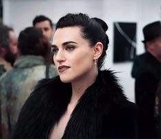 a woman with black hair wearing a fur coat and looking off into the distance while standing in front of other people