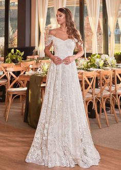 a woman standing in front of a table wearing a wedding dress with off the shoulder sleeves