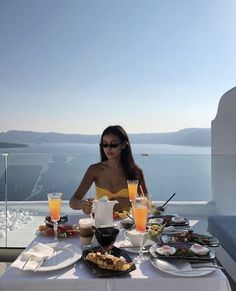 a woman sitting at a table with food and drinks in front of her, overlooking the ocean