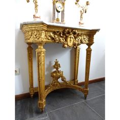 an ornate gold console table with statues on top and a clock in the corner next to it