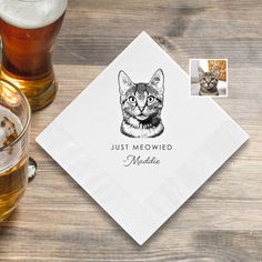 a napkin with a cat on it next to two mugs and a glass of beer