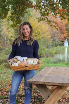 a woman standing in front of a picnic table holding a basket full of pumpkins