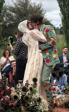 two people kissing in front of a group of people sitting on the ground and taking pictures