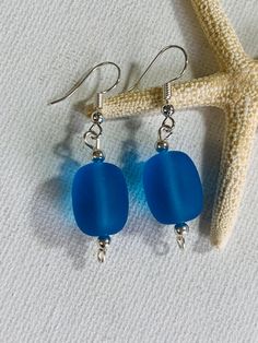 two blue glass beads hang from silver earwires next to a starfish on the beach