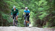 two bicyclists riding down a trail in the woods