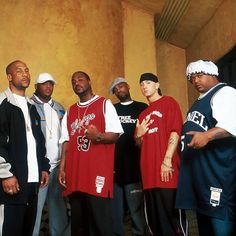 four men standing in front of a wall wearing red shirts and black pants with one man pointing to the side