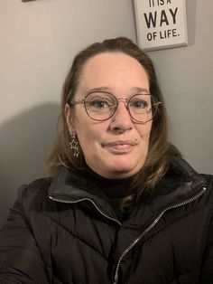 a woman wearing glasses and a black jacket in front of a sign that says it's a way of life