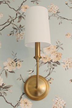 a gold wall light with a white shade on it's side against a floral wallpaper