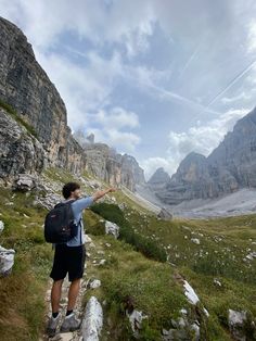 a man standing on top of a lush green hillside next to a rocky mountain range