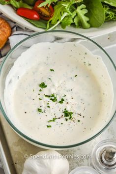 a glass bowl filled with white sauce next to a salad and bread on a tray