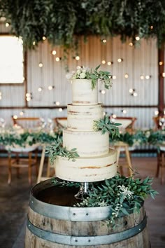 a wedding cake sitting on top of a wooden barrel in front of tables with greenery