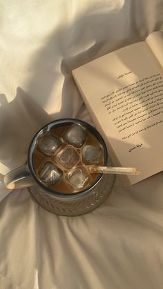 an open book sitting on top of a bed next to ice cubes in a cup