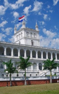 a large white building with palm trees in the foreground and a flag on top