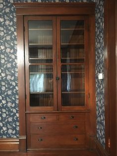 a wooden bookcase with glass doors and drawers in front of a wallpapered wall