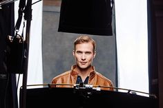 a man standing in front of a camera with his head turned to the side and wearing a brown leather jacket