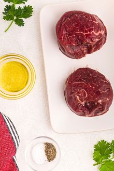 two raw meat patties sitting on top of a white plate next to some seasoning