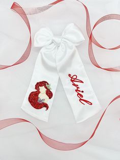 White satin bow with Ariel embroidery on it and a custom name. This bow also has glitter attached to it as well as crystals. Ariel Embroidery, Embroidery Hair Bow, Mermaid Bow, Embroidery Bow, Embroidery Hair, The Little Mermaid 2023, Little Mermaid 2023, Disney Bows, Sell Ideas