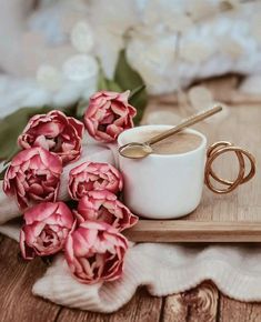 some pink flowers are sitting on a wooden tray with a cup of coffee and two gold spoons