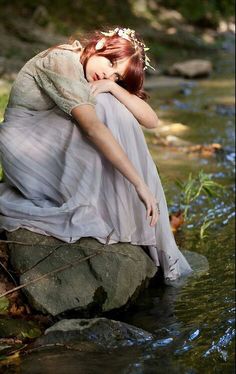 a woman sitting on top of a rock next to a river wearing a white dress