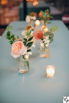 several vases with flowers and candles on a blue table cloth covered table in an indoor setting