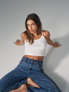 a woman in a white tank top and blue jeans sitting on a stool with her arms stretched out