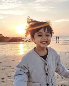 a young boy standing on top of a sandy beach next to the ocean at sunset