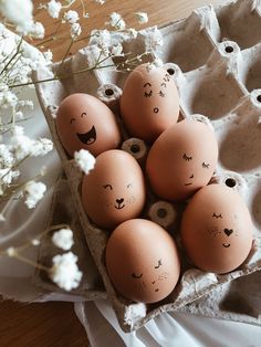 six eggs with faces drawn on them sitting in an egg carton next to flowers