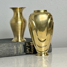 a gold vase sitting on top of a book next to a golden vase in front of a white wall