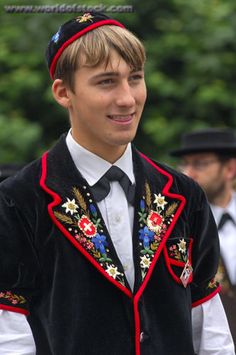 a young man wearing a black and red vest with embroidered flowers on the chest, standing in front of other men