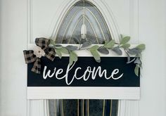 a welcome sign hanging on the front door with greenery and ribbon around it's edges