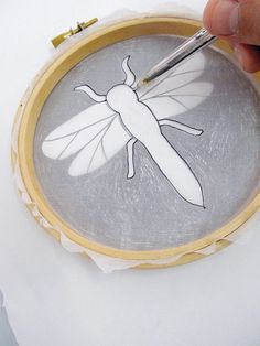 a person is using a needle to draw a bee on a piece of paper in a hoop