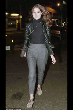 a woman in black jacket and grey pants walking down the street at night with her hand on her hip