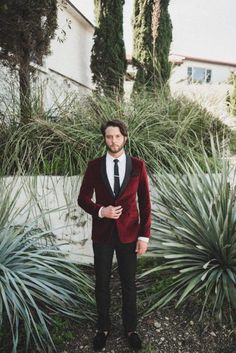 a man in a red suit and black tie standing next to some tall green plants