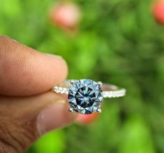 a person holding a ring with a blue diamond in it's middle and green plants in the background