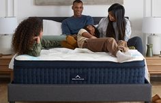 three people sitting on top of a blue mattress in a living room with white walls