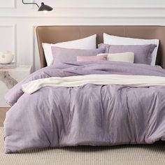 a bed with purple sheets and pillows in a white room next to a lamp on the wall