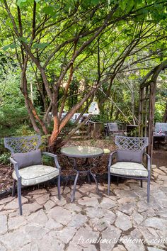 two chairs and a table in the middle of a stone patio under a large tree