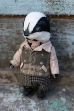 a small stuffed animal is wearing a jacket and hat, standing against a wooden wall