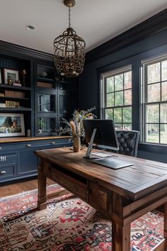 a desk with a laptop on top of it in front of two windows and a chandelier